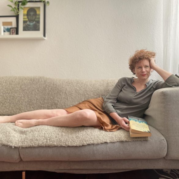 Auf dem Foto ist die Autorin Tina Molin zu sehen, die lächelnd und mit einem Buch in der Hand auf einem Sofa lehnt.